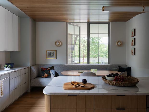 A refined open-plan kitchen and dining area with a large central island, integrated cabinetry, a built-in curved banquette, and a compact dining table. The room features warm timber ceiling boards and flooring, pale grey walls, light cabinetry, a pale stone island benchtop, and expansive black-framed windows overlooking greenery. Contemporary furnishings, framed artwork, and simple wall sconces create a calm, understated atmosphere.