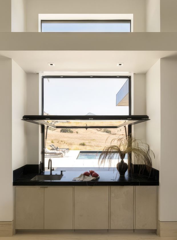 A compact contemporary kitchen wall with pale beige full-height cabinetry, a glossy black stone benchtop and matching dark splashback. A large open servery-style window connects visually to an outdoor pool and dry landscape, while a black horizontal awning or window element projects across the opening. Minimal decor includes a sink, tapware, fruit and a tall plant. A high-level window and recessed ceiling lights contribute to the bright, clean composition.