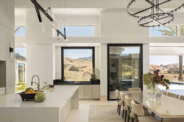 Bright, spacious open-plan kitchen and dining area with white minimalist cabinetry, a large island with an undermount sink and dark benchtop, and a long dining table with upholstered chairs. Black-framed glazing and doors open the room to a sunny landscape with rolling hills and palms. High ceilings, clerestory windows, and a sculptural black ring chandelier create a refined contemporary interior.
