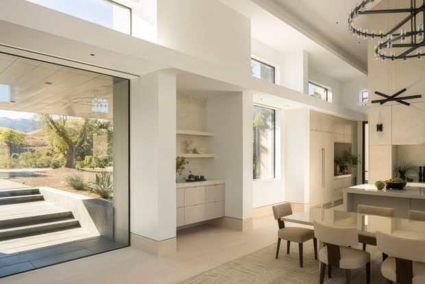 Bright, spacious contemporary open-plan kitchen and dining area with double-height ceilings, extensive floor-to-ceiling glazing, and a large glazed opening to a landscaped exterior. White walls, pale flooring, minimalist light cabinetry, integrated appliances, and a large dining table create an airy, refined interior. A sculptural black chandelier hangs above the dining area, while built-in shelving and cabinetry occupy the central wall.