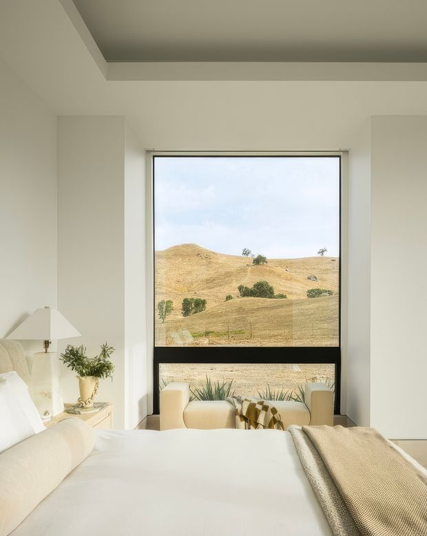 A bright, serene bedroom with a large black-framed picture window overlooking dry rolling hills. The room has white walls and bedding, a pale upholstered bed, light-coloured armchairs positioned by the window, a neutral throw blanket, a small bedside table with a lamp and potted greenery, and a recessed tray ceiling.