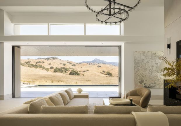 A bright, refined living room with a large open glazed wall framing a dry rolling rural landscape. A cream sectional sofa and sculptural lounge chairs surround a low coffee table, while a linear fireplace and large neutral-toned artwork occupy the right wall. High clerestory windows, white walls, pale timber ceiling lining, and a prominent dark metal ring chandelier define the spacious contemporary interior.