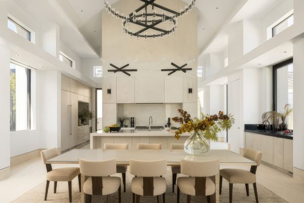 Bright, spacious open-plan kitchen and dining area with a dramatic double-height ceiling. The room features white built-in cabinetry, a large central island, a long dining table with upholstered chairs, extensive clerestory and full-height glazing, and distinctive contemporary black pendant lighting. The palette is light and neutral with warm timber flooring and subtle beige furnishings.