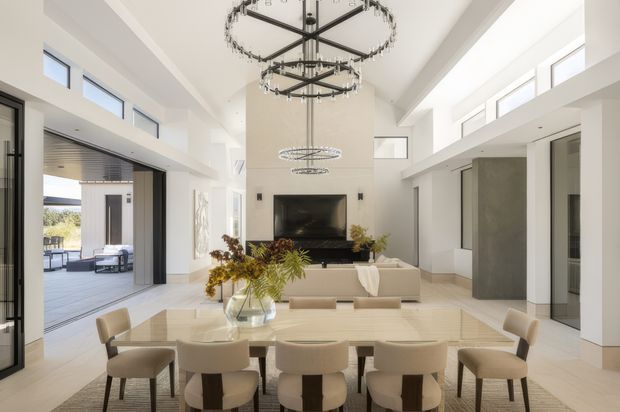 Bright, spacious open-plan living and dining area with a double-height ceiling, large windows and glazed doors opening to an outdoor terrace. A long dining table occupies the foreground, while a neutral sectional sofa faces a fireplace feature wall. Contemporary circular statement chandeliers, pale finishes and abundant daylight create an airy, refined interior.
