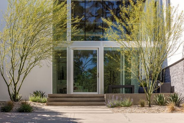 Front exterior view of a contemporary multi-storey building, featuring a tall glazed entrance framed in light-colored materials, dark upper-level glazing, concrete steps, and a minimalist xeriscaped garden with slender trees, succulents, gravel, and paving.