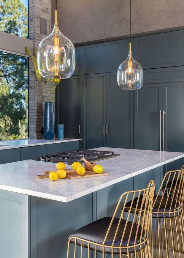 A refined contemporary kitchen centred on a large island with a pale veined stone benchtop, integrated gas cooktop, dark blue-grey panelled cabinetry and tall matching appliance cabinetry. Two clear glass pendant lights with exposed warm bulbs hang above the island, while gold-toned wire bar stools with dark seats provide contrast. Large windows bring in views of trees, and a tall blue vase adds a small decorative accent.