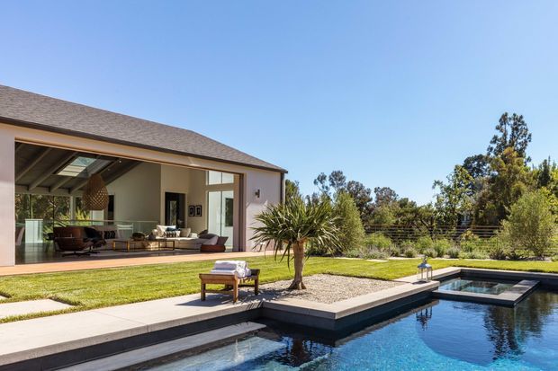 Contemporary single-storey residence viewed from the landscaped backyard, with a broad low-pitched roof, large glazed openings, and an open-plan living area extending onto a paved terrace. The foreground features a geometric swimming pool with raised rectangular sections, lawn, pale paving, gravel landscaping, and mature planting under a clear blue sky.