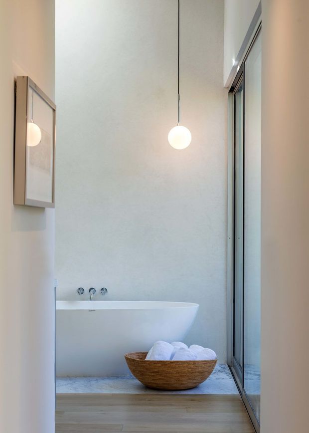 A serene, minimalist bathroom with a freestanding white bathtub positioned against a pale textured wall. A single spherical pendant light hangs above the bath, while full-height glass glazing or a glass shower enclosure lines the right side. The room features pale timber flooring, light stone or marble-look flooring around the tub, chrome wall-mounted tapware, and a simple framed wall mirror or cabinet. A natural wood bowl with folded white towels adds warmth.