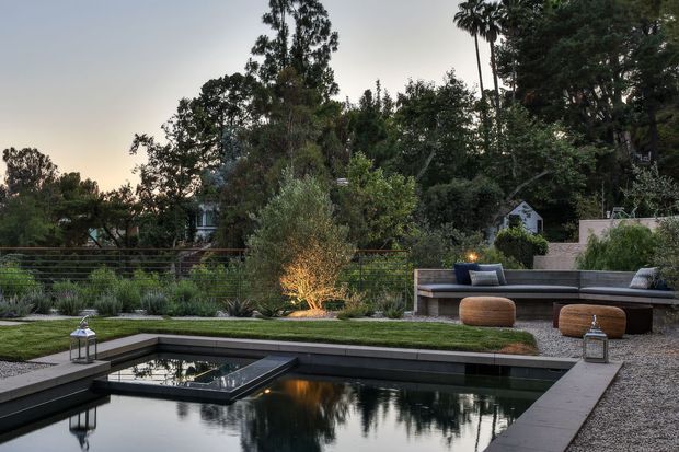 A landscaped outdoor courtyard at dusk featuring a long rectangular reflecting pool with dark stone or concrete edging, manicured lawn, layered planting, and a built-in outdoor lounge with a low bench, cushions, and woven ottomans. Lanterns and warm landscape lighting create a calm, refined atmosphere, with mature trees forming a private backdrop.