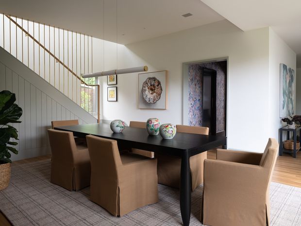 A refined dining room with a large dark timber dining table surrounded by upholstered tan dining chairs. The room features a sculptural pendant light, decorative ceramic vessels, artwork, pale walls, timber flooring, and a carpeted dining area. A stairway with slim vertical balusters and painted wall panelling is visible in the background, along with a dark-trimmed doorway and a glimpse of an adjacent room.
