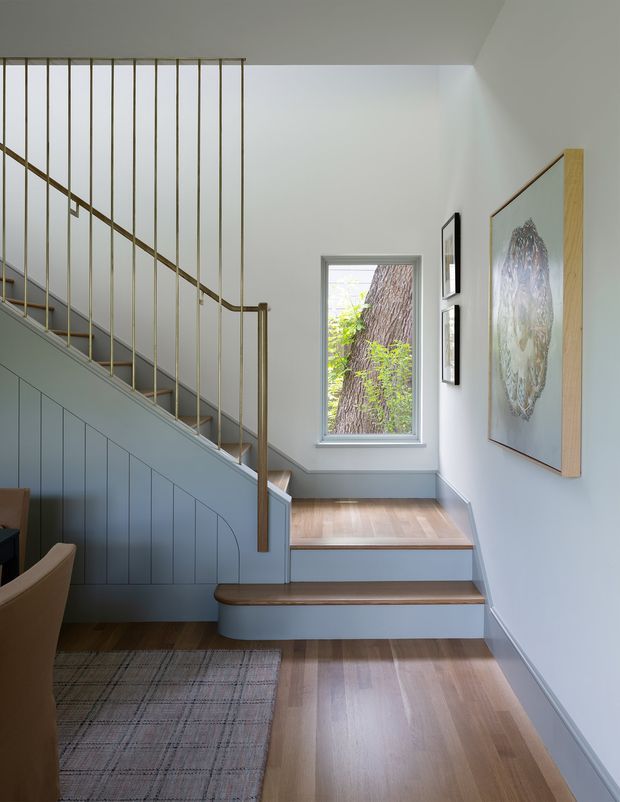 A refined interior view of a compact stairway and landing. The staircase has blue-grey painted panelled risers and side panelling, timber stair treads, and slender brass-toned vertical balusters with a matching handrail. Pale walls, timber flooring, framed artwork, and a narrow window overlooking a tree create a calm, light-filled setting. A portion of an adjacent seating area and rug is visible at lower left.
