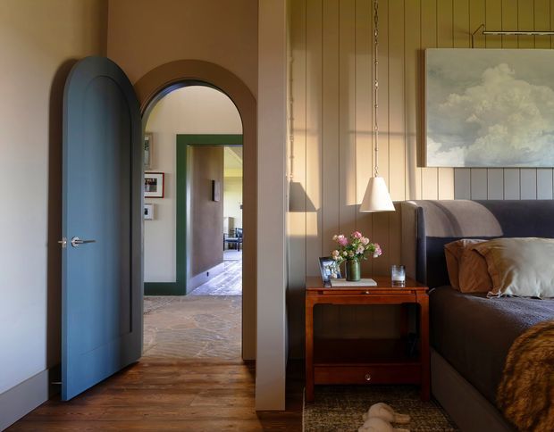Warm, refined bedroom interior viewed toward an arched doorway and hall. The room features vertical timber wall panelling, a dark upholstered bed with layered neutral bedding, a mid-century-style timber bedside table, pendant lighting, hardwood flooring, and a framed cloud artwork. A blue-grey paneled door and green-trimmed opening add colour and character, while the adjoining hall shows stone or patterned flooring.