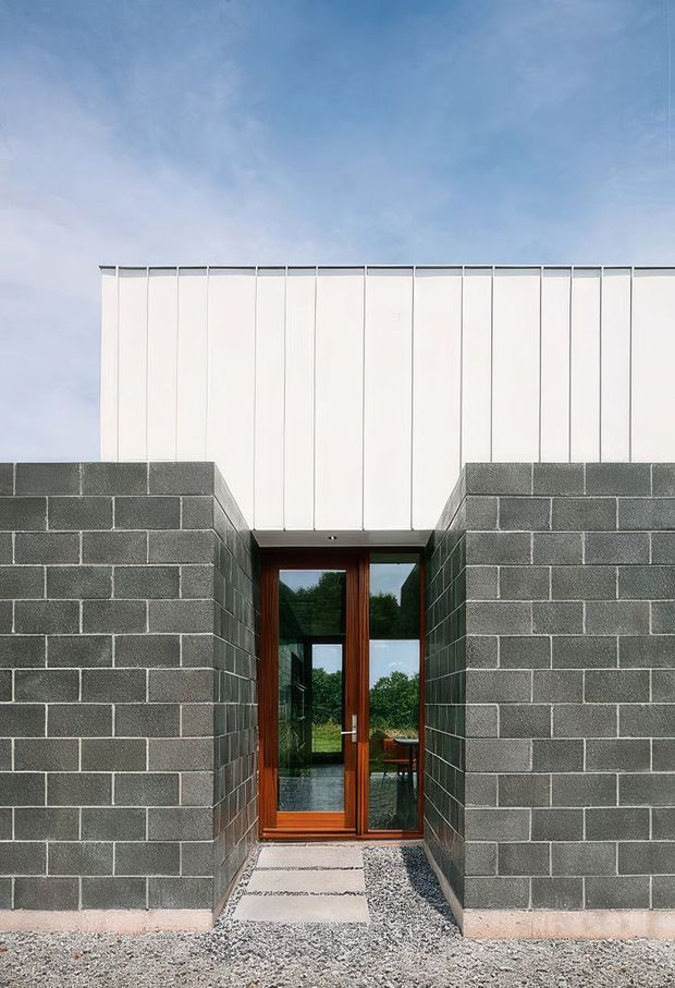 Front exterior view of a minimalist contemporary building with a narrow recessed entry framed by dark grey concrete block walls. A warm timber-framed glazed door sits beneath a tall white vertically panelled upper facade, with a gravel approach and simple concrete stepping path. The composition is geometric and restrained, with some greenery visible through the glazing.