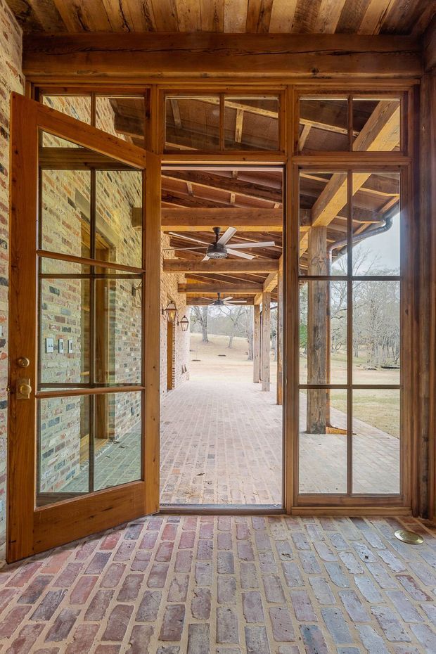 View from a rustic enclosed entryway through a timber-framed glazed door and sidelights onto a covered brick verandah. The space features exposed timber ceiling boards and beams, warm timber window and door frames, exposed brick walls, and brick paving extending from the interior to the outdoor porch. A ceiling fan and rural wooded landscape are visible beyond.