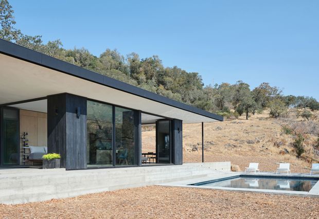A minimalist single-storey modern home set against a dry, wooded hillside. The structure has a long flat roof with dark metal edging, extensive floor-to-ceiling glazing, dark timber or charcoal-clad wall sections, and exposed concrete retaining walls. A narrow swimming pool sits beside the house, with a simple deck and loungers. The scene has a restrained indoor-outdoor composition and strong connection to the surrounding landscape.
