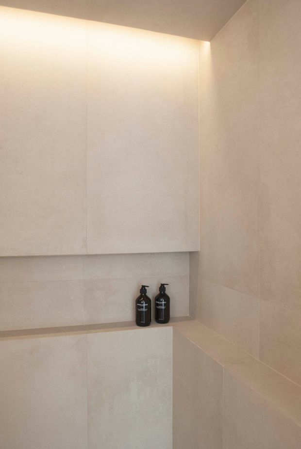 Minimalist close-up of a pale tiled bathroom corner with a recessed wall cabinet, integrated ledge, and two dark soap dispensers. Soft concealed lighting creates a warm, understated atmosphere.