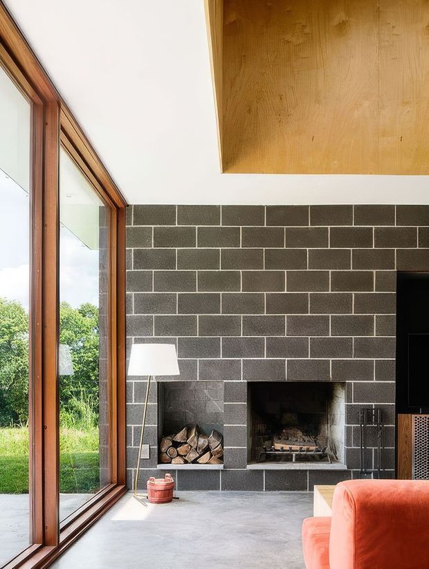 A bright contemporary living room with a dramatic dark grey masonry fireplace wall, exposed firewood storage, polished concrete floor, and a large timber-framed glazed opening overlooking greenery. A warm timber ceiling feature and orange upholstered seating add contrast to the restrained architectural palette.