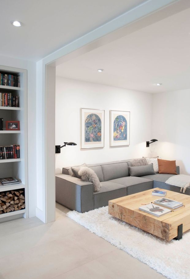 Bright contemporary living room viewed through a wide opening from an adjacent space. The room features a large grey sectional sofa, a chunky natural timber coffee table, a white shag rug, framed artwork, black wall-mounted reading lights, and recessed ceiling lighting. A built-in bookshelf with books and stacked firewood is visible at the left.