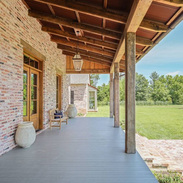 A covered outdoor veranda runs along a rustic brick building, with a deep timber-framed roof and exposed beams supported by substantial timber posts. The porch has a grey-painted decking floor, timber-framed glazed doors, a hanging lantern, ceramic urns, and a small wooden chair. Brick paving steps lead down to a broad lawn and landscaped grounds, with a small brick outbuilding visible beyond.