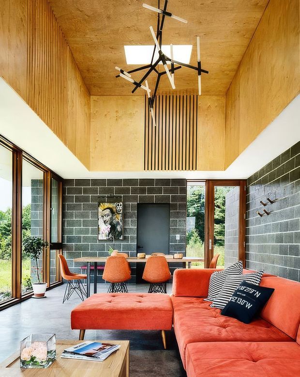 A bright, compact contemporary living and dining area with a vivid orange-red sectional sofa in the foreground and a small dining table with orange chairs behind it. The room features dark charcoal masonry block walls, extensive timber-lined ceiling and wall panels, full-height timber-framed glazing, polished concrete flooring, and a sculptural black-and-white pendant light. A garden is visible through the glazing.