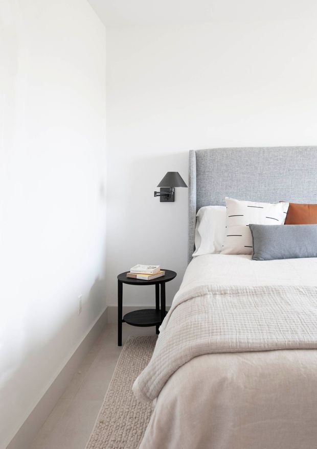 A bright, minimalist bedroom corner with a neatly made bed, a tall grey upholstered headboard, layered white, grey and terracotta cushions, and a textured light rug. A small round black bedside table with books sits beneath a dark wall-mounted lamp. The room has white walls, pale flooring and a calm, restrained palette.