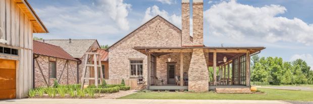 Wide exterior view of a low-rise brick residence or converted rural-style building complex, featuring gabled roof forms, a prominent tall brick chimney, timber-framed glazed outdoor living area, covered verandah, and landscaped garden beds. The composition has a warm, rustic character with clear farmhouse and barn-like influences.