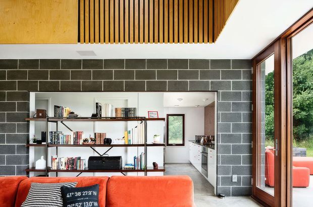 Contemporary open-plan living room with a vivid orange sectional sofa in the foreground, exposed dark concrete block walls, and black metal open shelving filled with books and objects. A compact white kitchen is visible through the opening, while full-height timber-framed glazing opens toward greenery outside. A slatted timber ceiling feature adds warmth and texture.