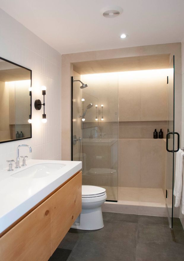 A contemporary bathroom with a large timber vanity and white integrated basin in the foreground, a black-framed wall mirror, and wall-mounted black-and-brass sconces. The rear of the room contains a glass-enclosed shower with beige tiled walls, recessed shelving, and a toilet. Large dark grey floor tiles provide contrast against the light walls and warm timber joinery.