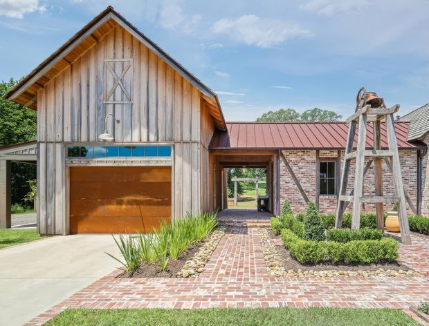 Exterior view of a rustic contemporary residential property combining a tall timber-clad gabled structure with an attached red-brick wing. The facade features a large timber garage door, blue clerestory-style glazing, exposed timber detailing, red metal roofing, and a brick-paved entry path bordered by landscaped planting and gravel. A timber garden structure is visible in the foreground.
