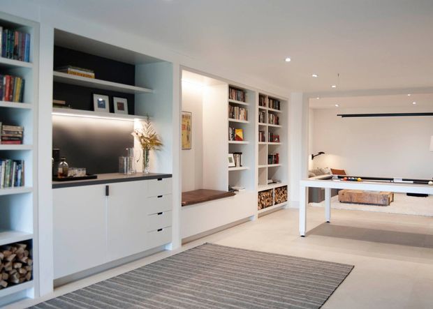 Bright contemporary open-plan recreation and living area with extensive white built-in shelving and cabinetry, integrated display niches, a dark benchtop, timber-lined storage cubbies, and a central timber bench seat. A full-size table-tennis table occupies the adjoining area, with a neutral striped rug, recessed ceiling lights, and warm under-shelf illumination.