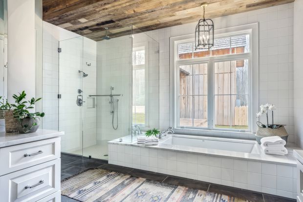 Bright, spacious bathroom featuring a tiled freestanding bathtub beneath a timber-framed window, a large frameless glass walk-in shower, white tiled walls, dark tiled flooring, white vanity cabinetry, and a rustic reclaimed-wood ceiling. A black cage pendant light, indoor plants, and folded towels add character.