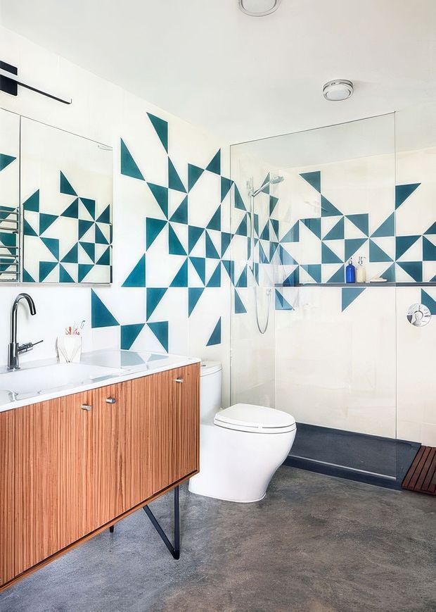A bright contemporary bathroom featuring a floating timber vanity with a white countertop and integrated basin, black tapware, a white toilet, and a walk-in shower with clear glass. The walls are decorated with a bold geometric pattern of teal and white triangular tiles, while the floor is a dark grey concrete-like finish. Large mirrors amplify the geometric wall design and reflect the shower area.