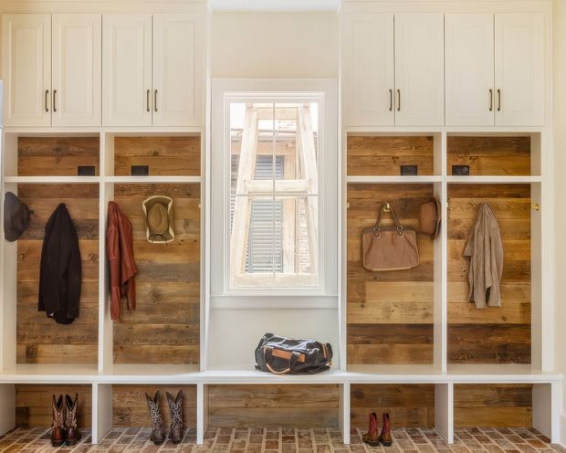 Built-in mudroom-style entryway storage fills both sides of a central window. White shaker cabinetry sits above open cubbies lined with reclaimed-looking timber, with hooks holding coats, scarves, hats, handbags and boots. A long white built-in bench and lower shoe compartments run across the bottom, set over a patterned brick or tile floor. The room has a warm, practical, traditional farmhouse-inspired character with abundant integrated storage.