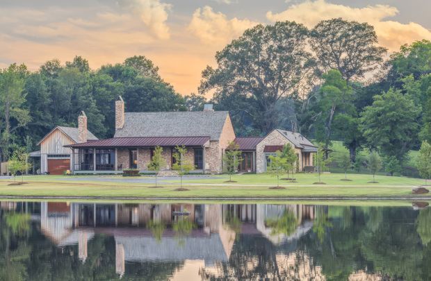 A broad exterior view of a substantial rustic country-style residence set beside a still pond, with multiple connected pitched-roof volumes, stone chimneys, timber and stone walls, and a covered verandah or porch. Mature trees frame the property and the building is reflected in the water. The warm evening light and landscaped lawn create a serene, rural atmosphere.