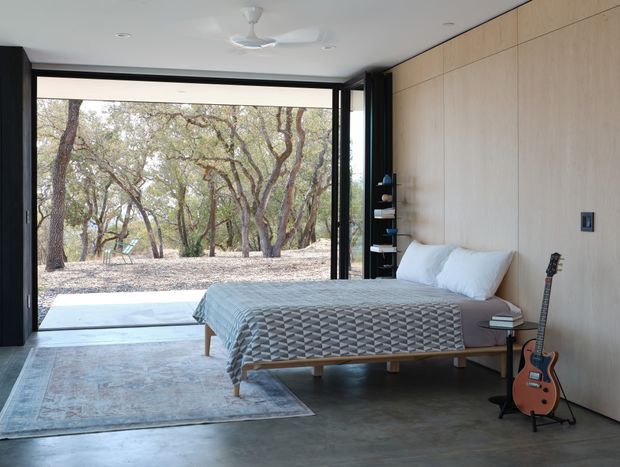 A minimalist bedroom with a low-profile bed positioned against a full-height light timber panelled wall. Black-framed floor-to-ceiling glazing opens directly to a wooded landscape, while polished concrete flooring, a large muted rug, recessed ceiling lights, a ceiling fan, and built-in open shelving create a clean, contemporary retreat. A guitar rests beside the bed.