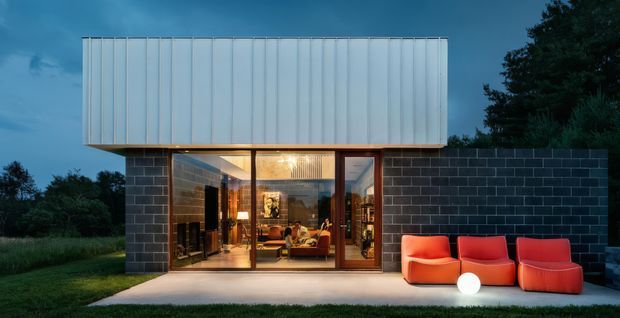 Twilight exterior view of a compact modern house with a rectilinear upper volume clad in pale vertical metal panels, a dark masonry lower wall, and a fully glazed living area opening onto a concrete patio. Bright orange-red outdoor lounge chairs and a glowing spherical light sit in front of the glazing, with trees and lawn surrounding the building.