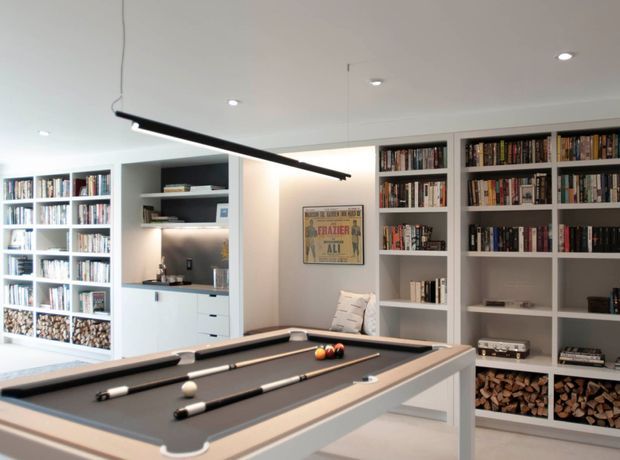A bright contemporary recreation room centred on a full-size pool table, with extensive built-in white shelving filled with books and firewood. Integrated cabinetry and a small illuminated display niche line the rear wall, while recessed ceiling lights and a long black linear pendant provide modern lighting. A framed poster adds a small accent to the otherwise white and grey interior.