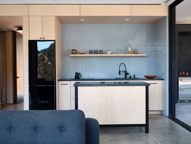 A contemporary open-plan kitchen viewed from the living area, featuring pale timber flat-panel cabinetry, a central island with a dark benchtop and black framed base, a pale blue-grey vertically tiled splashback, open timber shelving, and black tapware and appliances. Full-height timber cabinetry surrounds the kitchen, with a glazed opening to an adjacent area and a blue upholstered sofa in the foreground.