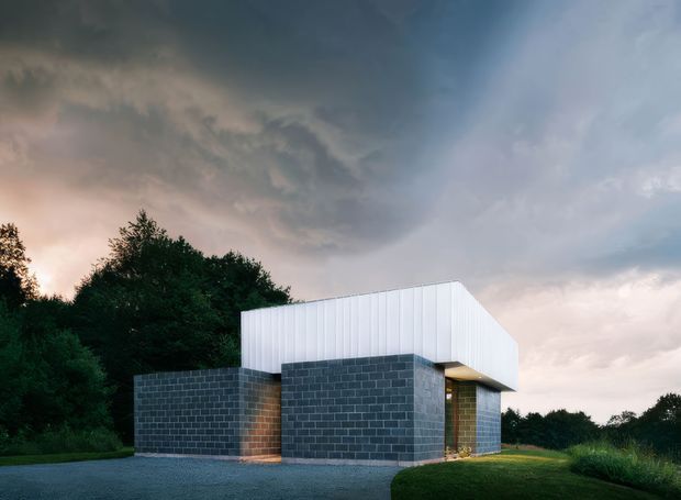 A compact contemporary flat-roof building set against dense trees and an open grassy landscape. The lower walls are constructed from dark grey concrete masonry blocks, while a crisp white vertically ribbed upper volume projects over the darker base. A recessed warm-lit entrance is visible beneath the overhang, with a paved forecourt in front. The dramatic cloudy sky creates a moody architectural exterior scene.