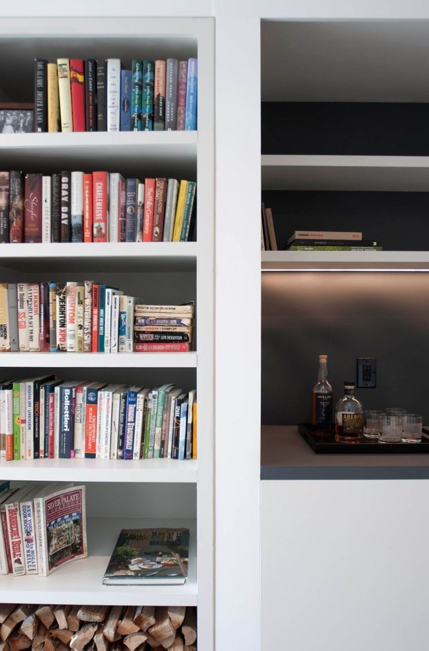 Close-up interior view of custom white built-in shelving. The left shelving is filled with books and magazines, while the right section forms a dark recessed display niche with integrated warm lighting, bottles, glasses, and a small electrical outlet. Firewood is stored in the lowest visible compartment. The joinery has a clean, contemporary built-in appearance with strong white-and-charcoal contrast.
