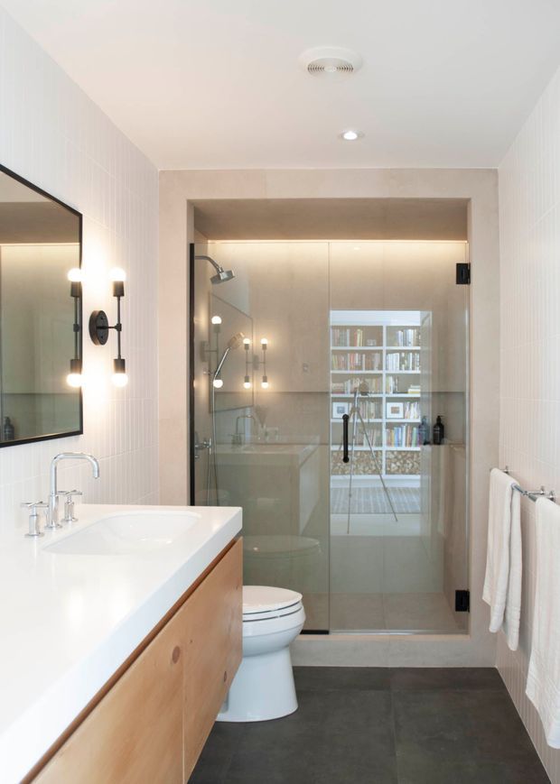 A narrow, contemporary bathroom with a wall-mounted timber vanity, white integrated basin, chrome tapware, large black-framed mirror, wall sconces, dark grey floor tiles, and white vertically textured wall tiles. A frameless glass shower enclosure occupies the rear, with a bathtub and shelving visible through reflections.