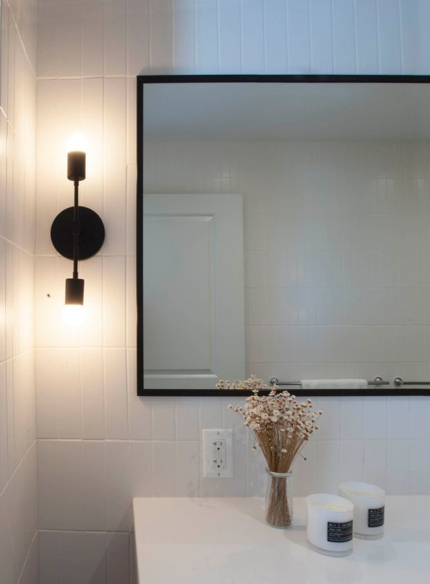 Close-up view of a bright bathroom vanity area with white square wall tiles, a large rectangular black-framed mirror, black double wall sconce, and a white countertop with decorative candles and dried flowers. The mirror reflects a white paneled door and tiled wall.
