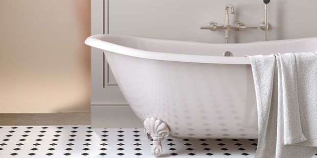 A close-up view of a freestanding white clawfoot bathtub in a refined bathroom. The tub has ornate white feet, exposed chrome plumbing and shower fittings, and a white towel draped over the rim. The room features pale blush-beige wall panelling and a black-and-white geometric tiled floor, creating a classic, elegant atmosphere.