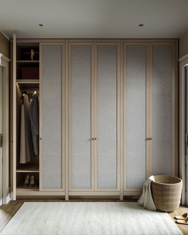 A compact, refined walk-in wardrobe with full-height built-in cabinetry across the back wall. The wardrobe doors have pale taupe timber frames and textured light fabric or mesh inset panels, with open shelving and hanging garments visible at the left. A soft neutral rug covers the timber floor, while a woven basket and draped textile add warmth near the doorway. Recessed ceiling lights and clean contemporary joinery create a calm, organised interior.
