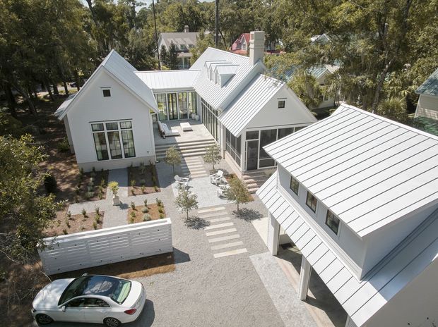 Aerial view of a contemporary white-clad residential compound surrounded by mature trees. Multiple pitched-roof gabled volumes with standing-seam metal roofing are arranged around a landscaped gravel courtyard with stepping-stone paths, outdoor seating, and a covered carport or parking structure. Large glazed openings connect the buildings to the central courtyard.