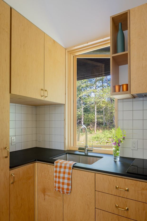 Compact L-shaped kitchen with warm natural wood cabinetry, black countertops, a white square-tile backsplash, and brass hardware. A large timber-framed window above the sink provides views of surrounding trees, while open shelving displays simple ceramic vessels. The space is tidy, bright, and functional with a restrained mid-century-inspired character.