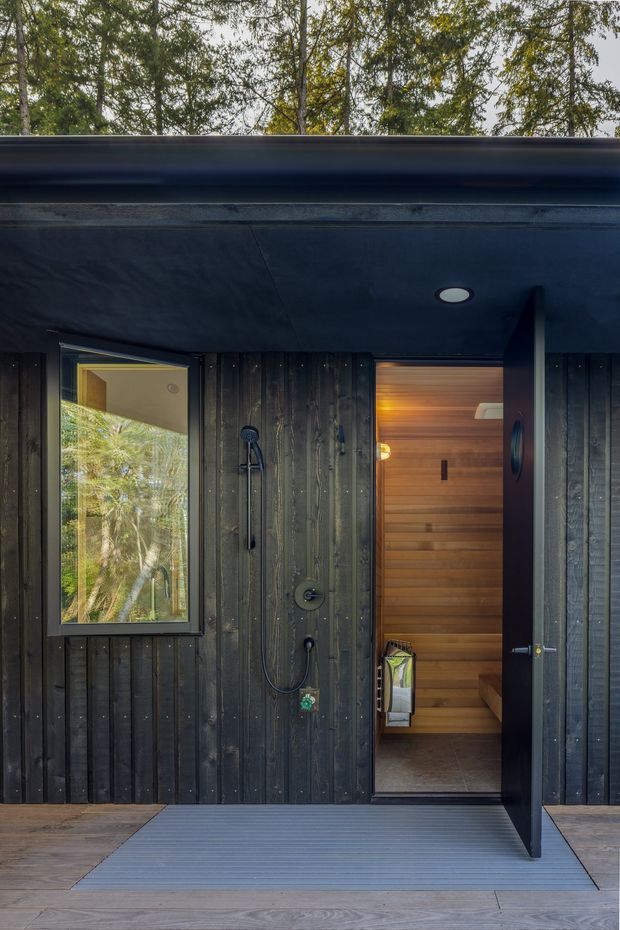 Exterior view of a dark timber-clad wet-room and sauna entry opening onto a timber deck. The black vertical cladding includes an outdoor shower beside a glazed window, while the open dark door reveals a warm timber-lined sauna with built-in bench seating. A recessed ceiling light and surrounding trees complete the forested setting.