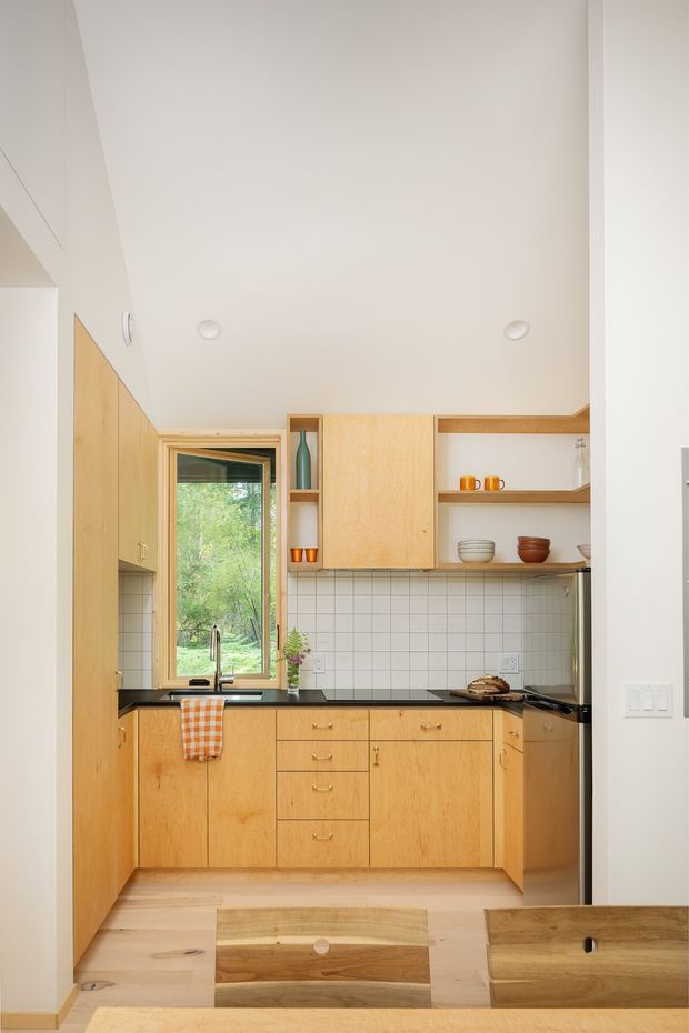 A compact, bright galley-style kitchen viewed from an adjoining dining area. The room features warm natural timber cabinetry, a black benchtop, white square tiled splashback, open timber shelving, a stainless-steel sink and tap, integrated appliances, and a timber-framed window overlooking greenery. A timber dining table and chairs are visible in the foreground.