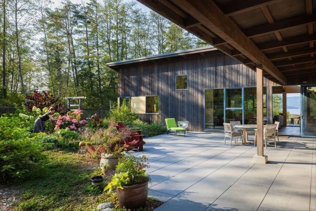A covered outdoor terrace beside a modern timber-clad house, with a large paved patio, timber posts and exposed roof structure, outdoor dining furniture, and landscaped garden planting. The house has dark vertical wood cladding and extensive glazing, with a glimpse of water or landscape beyond.