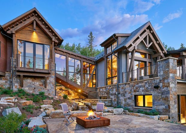 Exterior view of a substantial multi-level mountain-style residence built into a rocky landscaped site. The home combines extensive natural stone masonry, dark timber framing and gabled roofs with large glazed openings and balconies. A paved terrace with a central outdoor fire pit and lounge chairs occupies the foreground, while warm interior lighting creates an inviting evening atmosphere.